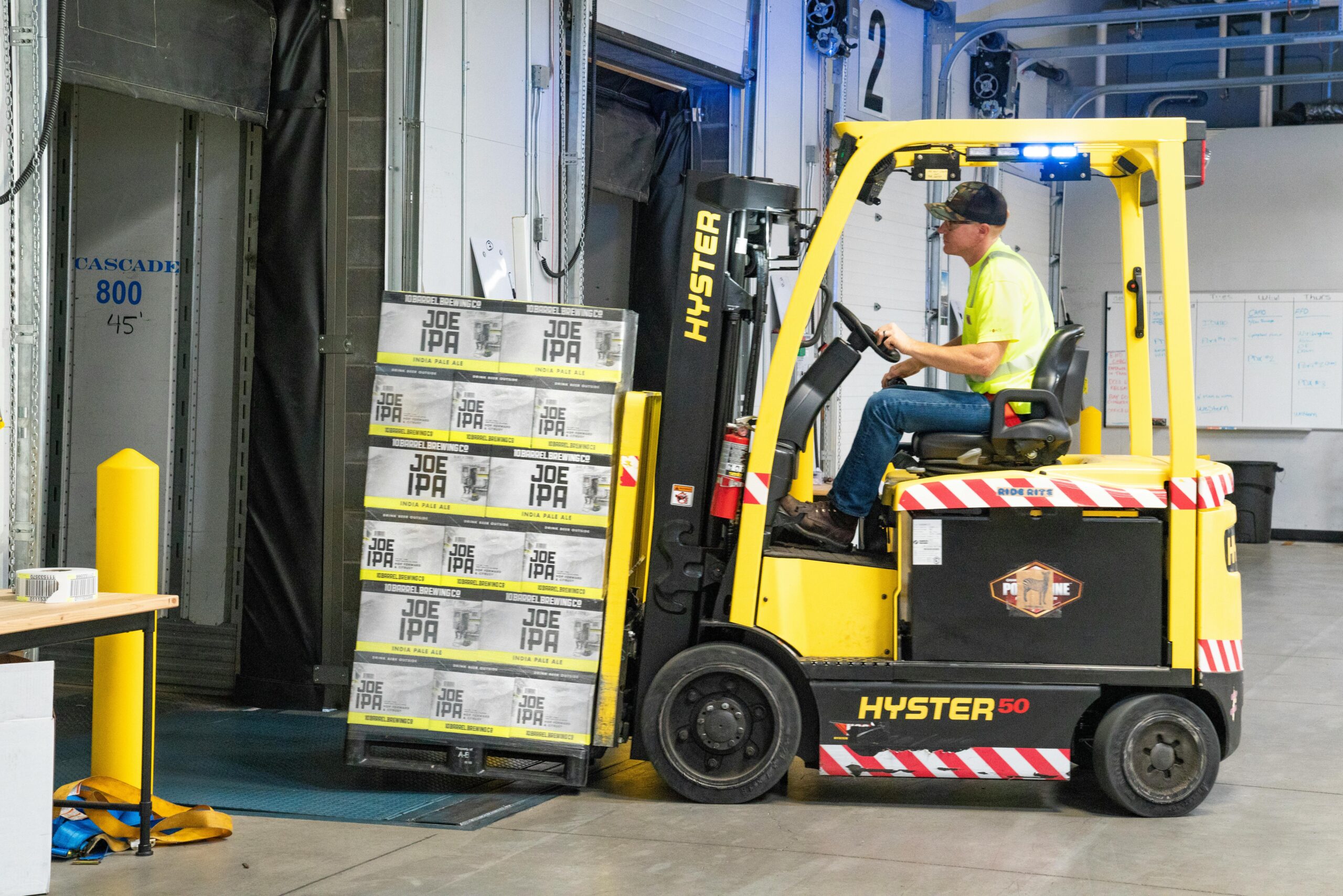 A worker drives a Hyster forklift moving Joe IPA boxes in a warehouse.